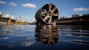 Construction site for an immersed tunnel beside a wide port channel, with concrete tunnel sections, cranes, boats, and city buildings in the background.